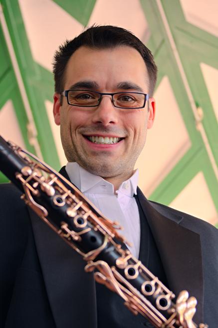 Matthew Boyles against a green and white pattern background, smiling down at camera, whilst nicely dressed and holding his clarinet